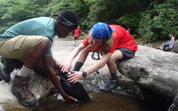 Two students kneel on rocks as they fill a water bladder in shallow water.